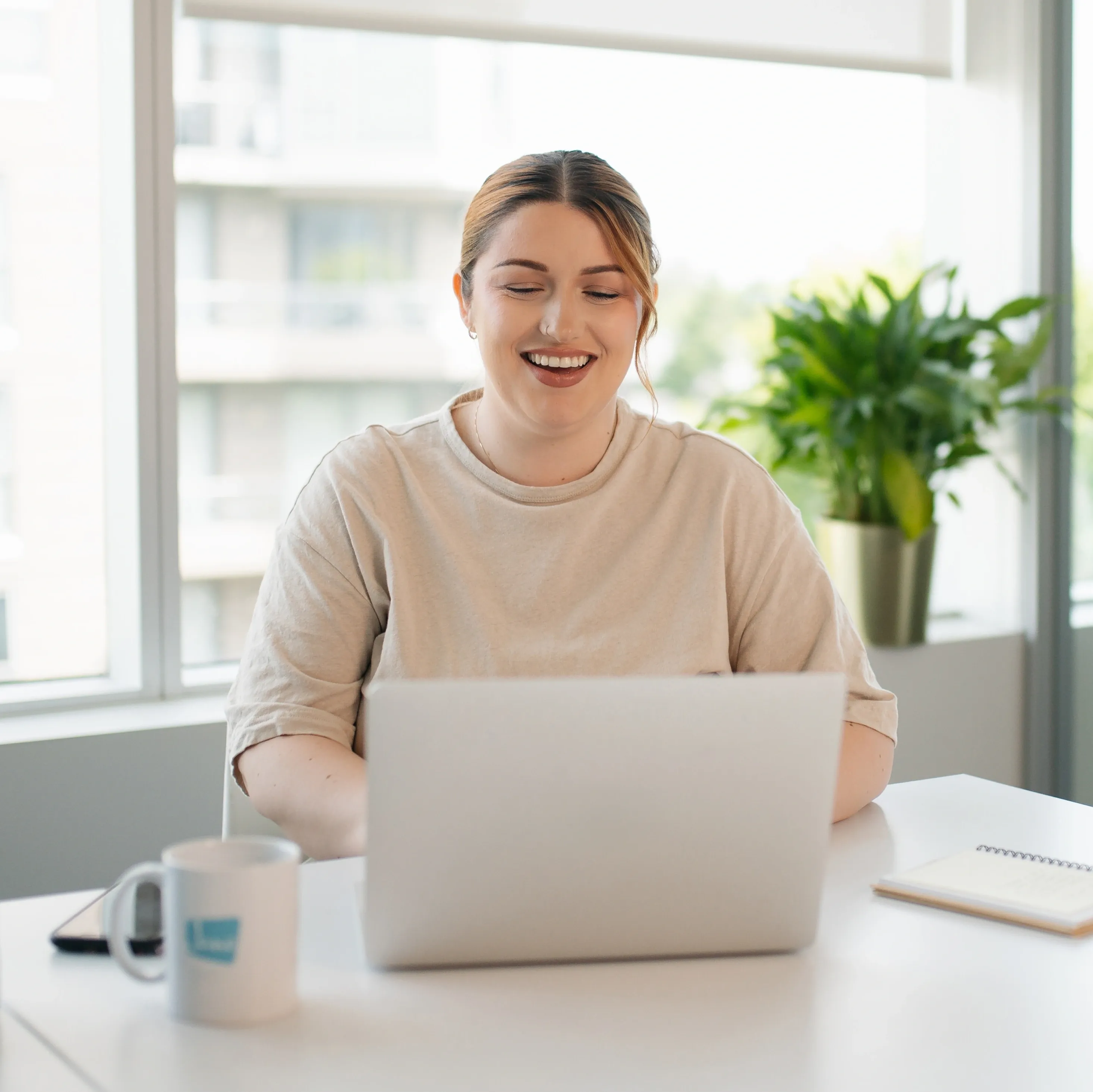 A smiling Jane team member works on a laptop in a bright, modern office with Jane-branded mug and water bottle on the desk.