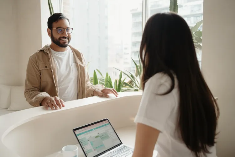 A patient smiles while checking in at a clinic front desk, where staff use Jane’s practice management software on a laptop.