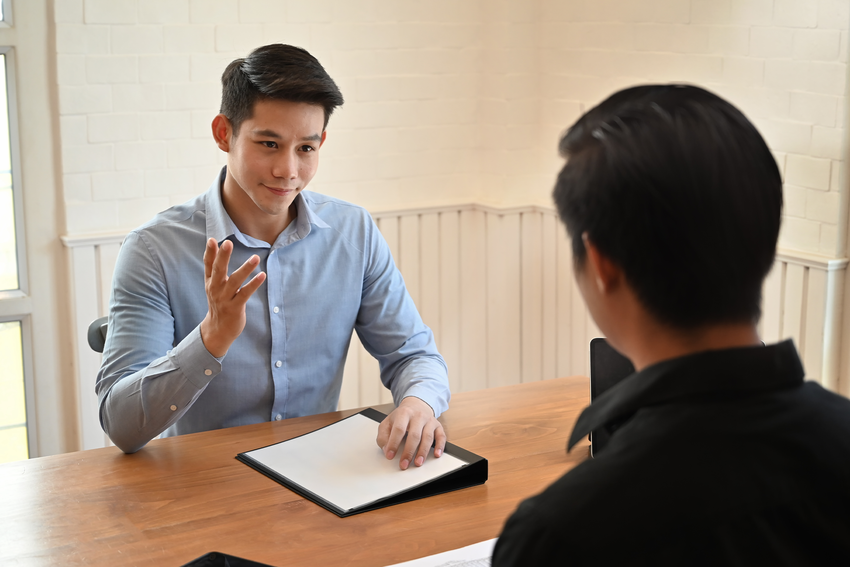 Two people sit at a desk in an office. One man wears a long blue dress shirt and has short dark hair. He holds his hand up as he listens to the person on the other side of the desk speak. His other hand is on top of a white sheet of paper on a black binder. The other person is a silhouette and we can only see behind them. They have short, dark hair.