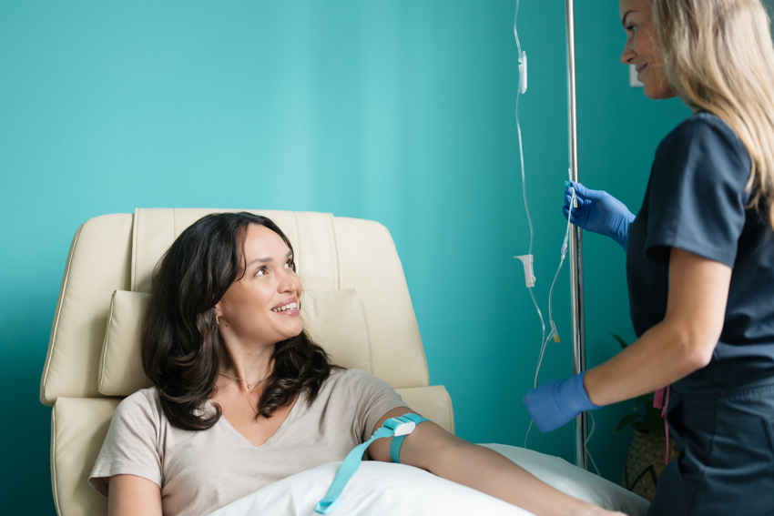 A patient sits in a treatment room chair and smiles at the practitioner standing next her. The patient has long dark hair and holds a pillow on their lap. A light blue strap is tightened around her arm. The practitioner wears dark blue scrubs and light blue gloves, and has long, blonde hair. She holds an IV stand. The background of the treatment room is light blue.