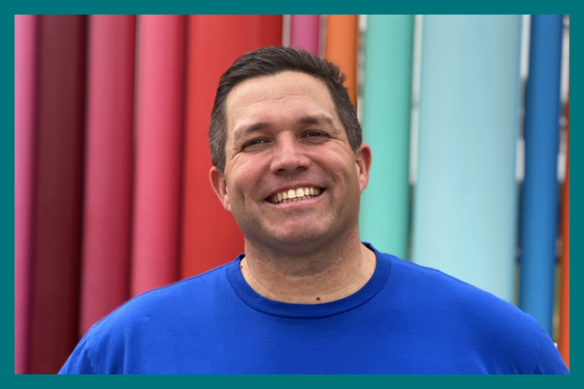 Dr. Josh Saterlee smiles for the camera. He wears a blue shirt and has short dark hair. His background is multi-colored panels that range from shades of purple and red to blue.