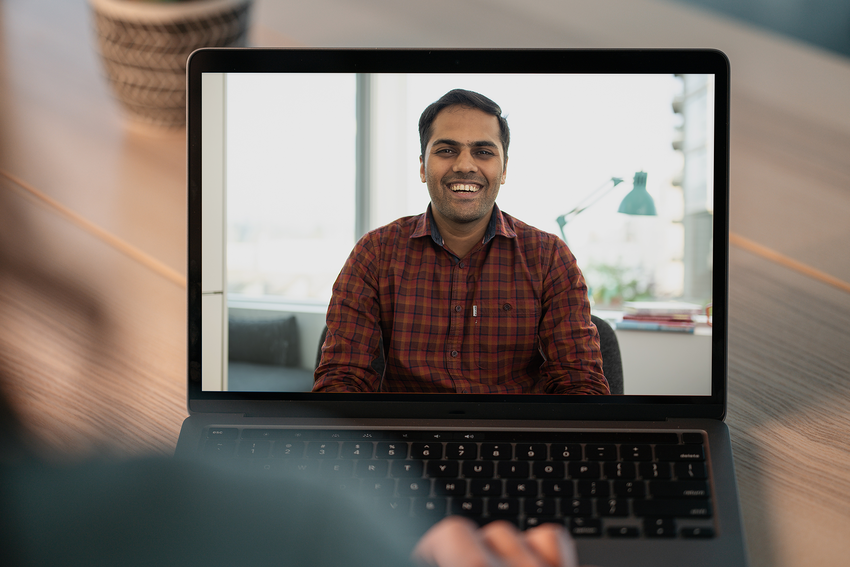 A therapist appears on a laptop screen. He smiles and wears a red plaid shirt. His black hair is short and we see a bit of his office behind him over the shoulder of the client, whose image is blurred.