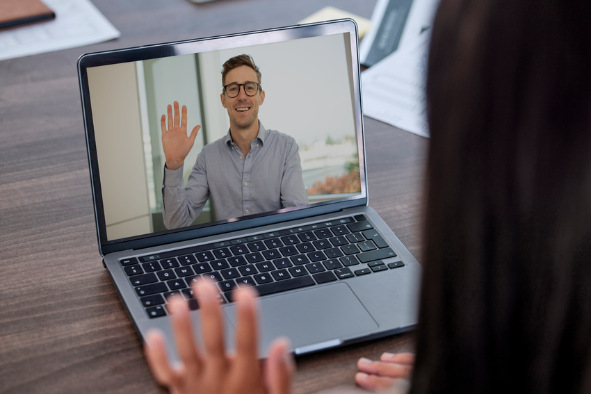 An over-the-shoulder view of an interviewee looking at their laptop on their desk and waving to an interview candidate on screen. The candidate is smiling. They have short brown hair and a blue dress shirt. The candidate waves to the interviewee. They are in a white office setting with a large window behind them, where there is the top of a tree visible.