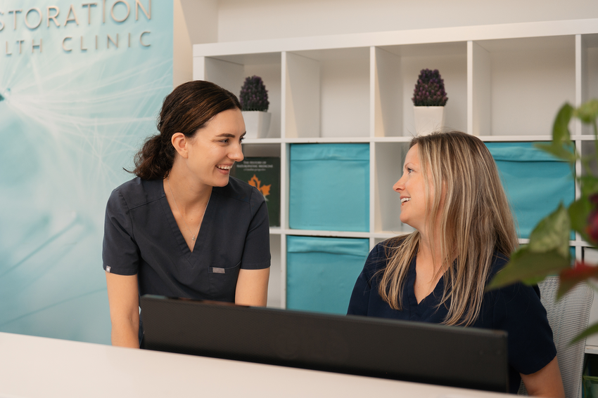 A clinic staff member smiles and chats with a front desk staff member. The clinic staff member has dark hair pulled back into a ponytail. The front desk staff member has long blonde hair. They both wear navy blue scrubs. Behind them, their clinic's name, Restoration Health Clinic, is on the wall in silver letters against a blue and white backdrop, and there is a white shelving/storage unit with blue bins and plants placed throughout the shelves.