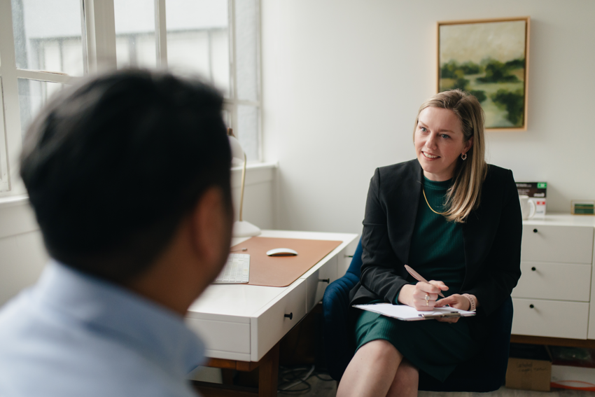 A practitioner practices active listening in conversation with their patient. The practitioner has long blonde hair and wears a dark green dress with a black blazer, complemented by a gold necklace and earrings. They have a pen in hand, notebook on their lap, and smile as their patient speaks. We only see the patient's silhouette from the back. The treatment room has white walls with a set of white dresser drawers behind the practitioner and a gold-framed painting of green hills and trees above that dresser. To the practitioner's right, there is a white desk with a corkboard pad and white mouse and keyboard. Further to the right, windows reveal soft grey light from outside.