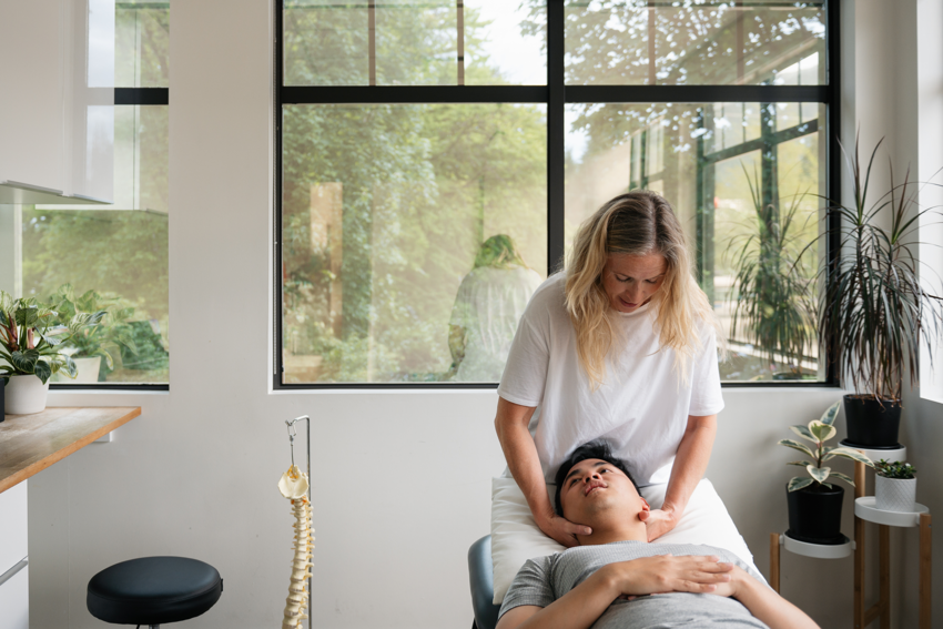 A patient lies on a bed in a treatment room. The practitioner speaks to the patient while cradling their neck. The patient is wearing a grey t-shirt, his arms crossed over his stomach. The patient has short, black hair. The practitioner has long blonde hair and wears a white t-shirt. The treatment room has a window that looks out into a forested area with lots of lush trees. Inside the treatment room, the walls are white and there are an assortment of plants in the right-hand corner behind the patient and practitioner. To their left, there is a model of a spine, a black stool, and a work table with cabinetry. On the table, there is a potted plant.