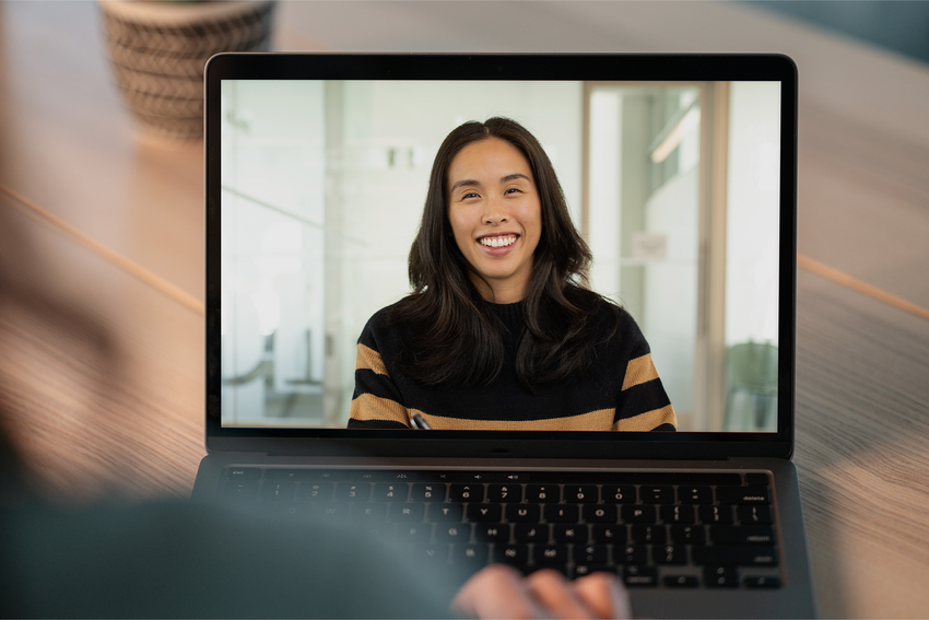 Someone at a desk looks at a laptop screen on a wooden desk. We can only see over the person's shoulder, which is blurred out. A woman with long dark hair appears on-screen. She smiles and wears a black and brown striped sweater. Her background is slightly blurred with glass walls and a wooden sliding door. She holds a pen.