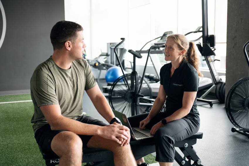 A practitioner speaks with her client in a gym setting in a clinic. The practitioner has long, red hair tied back into a ponytail and wears a black shirt and black pants. The patient has short brown hair and wears a green shirt and black shirt. Behind them, there is green astroturf and equipment like treadmills, yoga balls, and ellipticals.