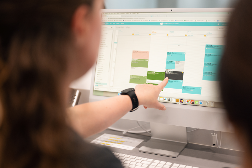 We see a desktop computer screen with Jane's EHR software open to a weekly schedule with various blocked times in various colors. Someone on the left points to the screen mid-conversation with someone on the right. We only see the silhouettes of these people from behind.
