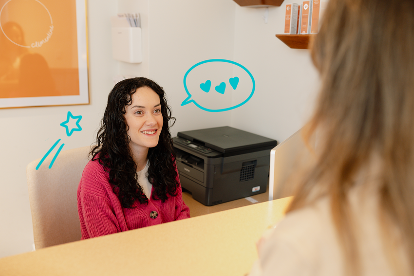 A front desk staff member greets a patient as they approach the desk. The staff member has long, dark curly hair and wears a white shirt with a pink cardigan on top. The clinic's walls behind the staff member feature orange products on shelves and a framed logo of the clinic's name, Clementine, in orange and white. A printer sits on the side of the desk. There are illustrated elements of a blue shooting star and a blue speech bubble with three hearts around the staff member.