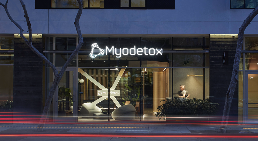 The exterior of Myodetox's clinic in West Hollywood, Los Angeles at night. We see the logo in white and can see into the reception with the front desk and a staff member standing at the desk.