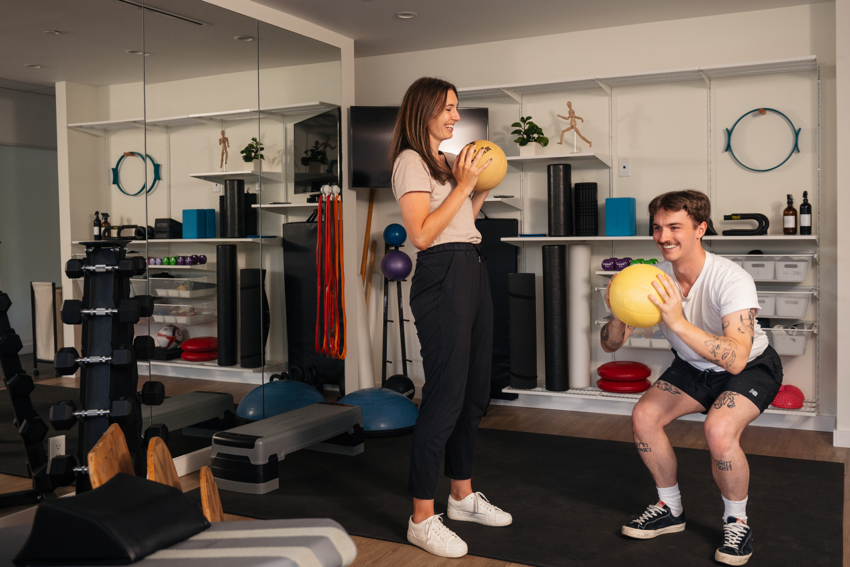 A gym room at a clinic. A practitioner instructs a patient on how to use a yellow medicine ball in the middle of an exercise. The practitioner has long brown hair and wears a beige shirt with black pants and white sneakers. The patient beside her is in squat position, with short brown hair, and is smiling. He wears a white t-shirt, black shorts, and black and white sneakers. He has an assortment of tattoos on his arms and legs. The gym has a mirrored wall to the left with weights, and the wall behind them has an assortment of other gym equipment, a plant, and a wooden model in a running position.