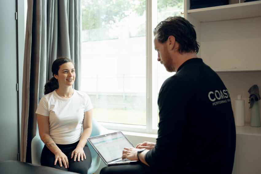 A patient and practitioner sit in a treatment room. The patient wears a white t-shirt and dark pants, and has long, dark hair pulled into a ponytail. She has a tattoo on her right arm. She smiles as she speaks to the practitioner. We see the practitioner from the back. He wears a dark long-sleeve shirt with the word "COAST" on the back. He has short dark hair and a beard. He is typing up notes on a laptop. The treatment room has grey walls, dark grey curtains, and white shelving. The patient and practitioner are in front of an open window with light streaming in and there are trees in the distance.
