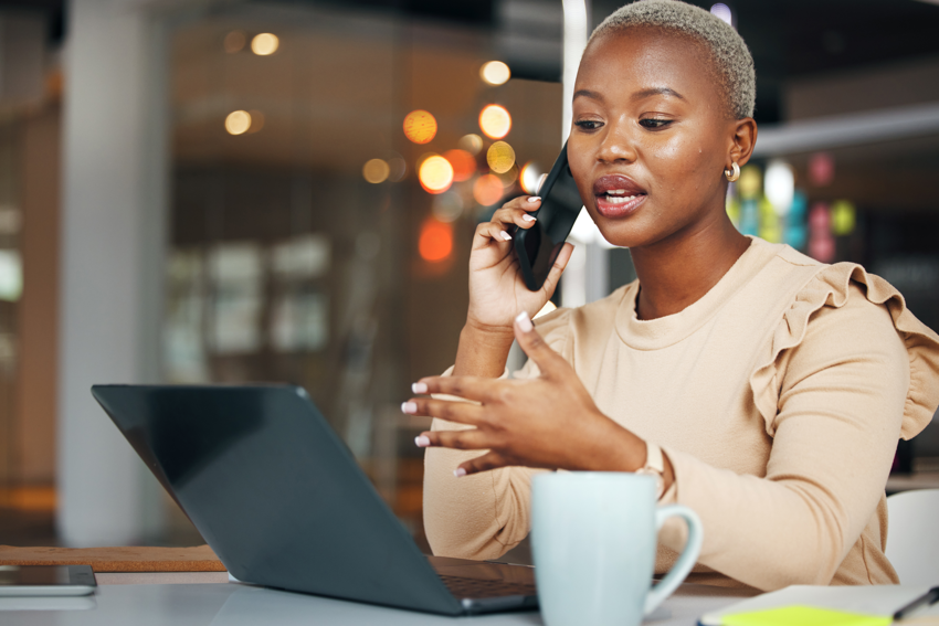 A woman talks on the phone while in front of her laptop in her office. She has short, blonde hair and wears a beige blouse.