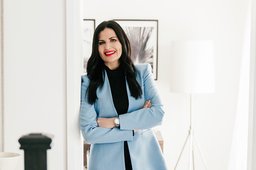 Nicole McCance smiles for the camera. She leans against an open doorway. The whole room in front of her and the room behind her are white. She wears a powder blue blazer with a black turtleneck and a silver watch. She has long, dark hair and red lipstick on. Behind Nicole, the room has black and white framed photos and a silver floor lamp with a white lampshade.