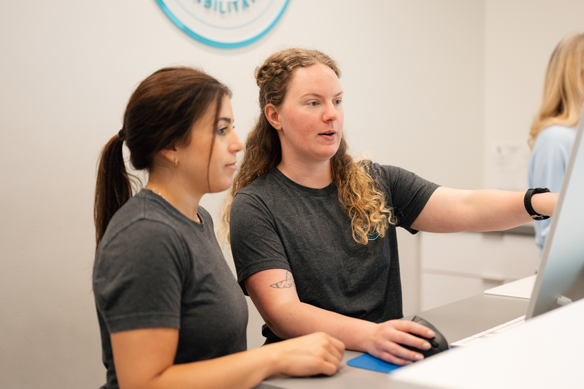A clinic staff member trains a front desk staff member. They both wear dark grey shirts. The trainer, who has long, curly blonde hair, points to a computer screen in front of them. The trainee, who has long, dark hair pulled back into a ponytail, looks at the screen. The clinic's walls are white and we see a bit of their light blue, circular logo behind them.