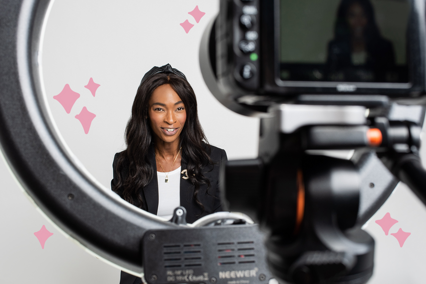 Lisa Simone Richards, PR and visibility strategist, smiles at a camera with a ring light attached to it. She wears a black headband, black blazer with a white shirt underneath, and a gold lock necklace. Illustrated pink stars float around her.