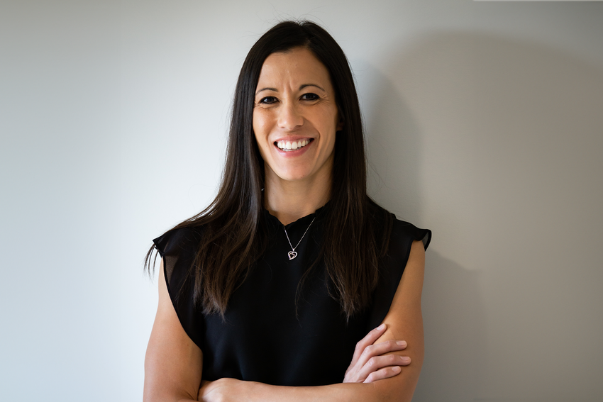 Stephanie Honess, a physiotherapist and the owner of [Waso Physiotherapy], smiles for the camera against a white backdrop. She has long dark hair, and a black top on with a silver heart pendant necklace. She crosses her arms.