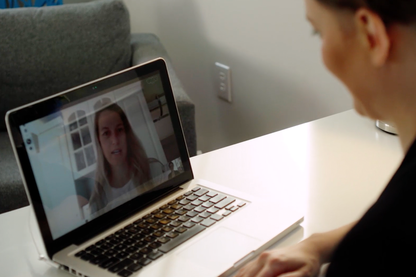 Julia Smith meets with a client virtually. We see behind Julia's shoulder that she is looking at the screen of her laptop with a blurred out view of a patient.