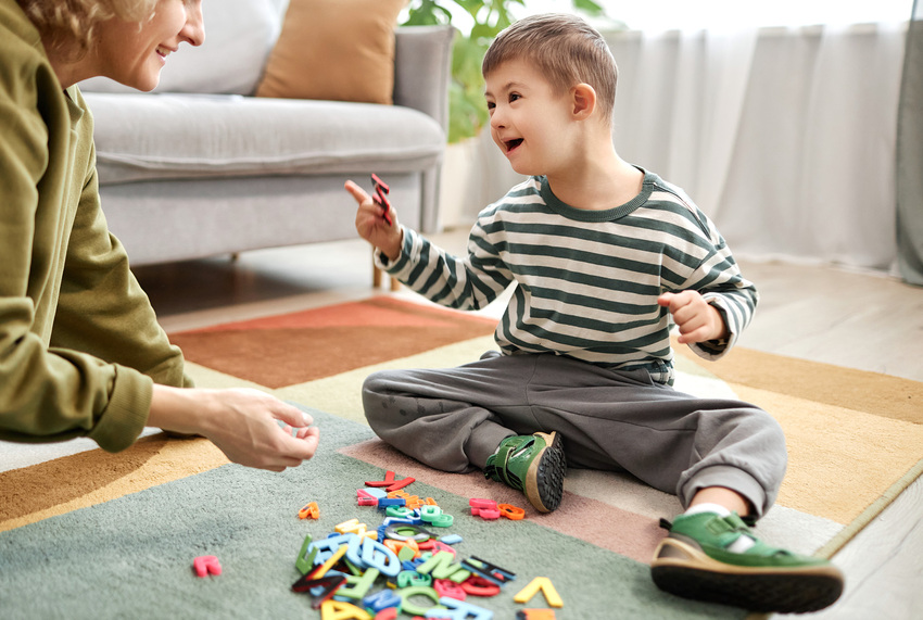 A young child smiles and plays with block letters on the play mat of a waiting room. Their parent or guardian is crouched beside them, smiling.