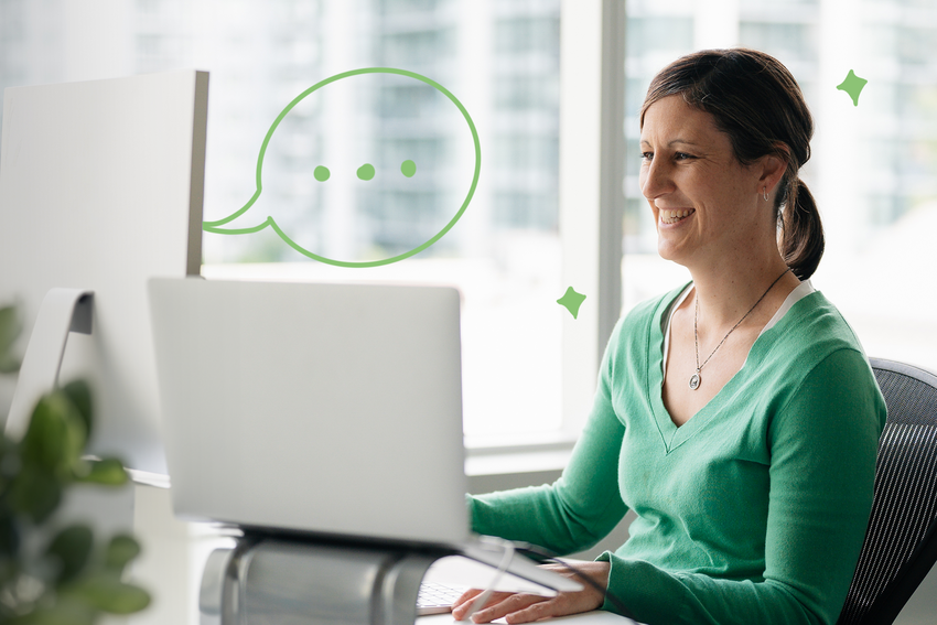A woman in an office looks at a desktop computer and smiles. She wears a long-sleeved green shirt and has a long necklace on. Her brown hair is tied back into a ponytail. There are illustrated elements of a speech bubble coming from the computer screen and little green stars around the woman.